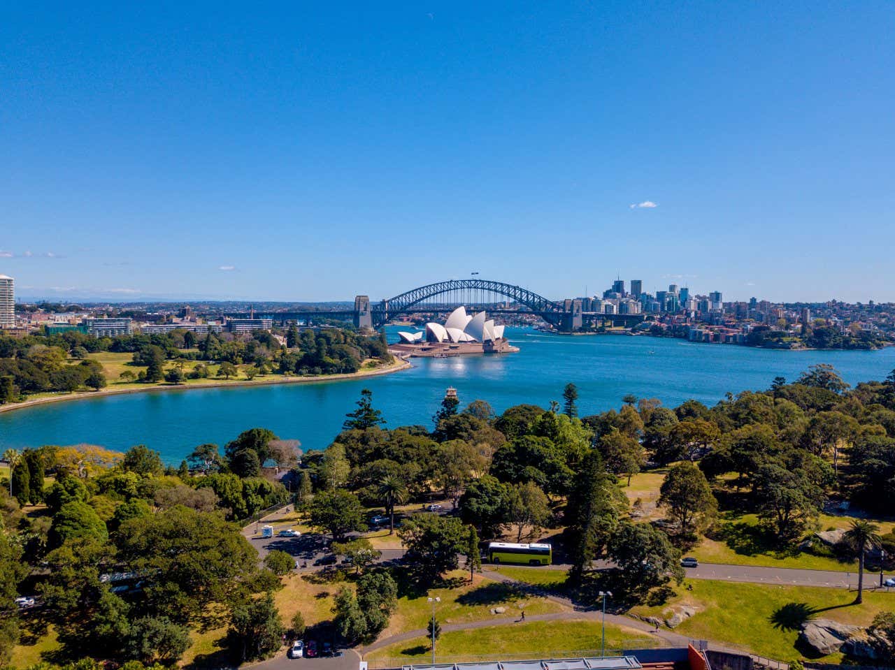 An aerial view of Sydney Opera House, seen from afar, with Harbour Bridge in the background, and a clear blue sky.