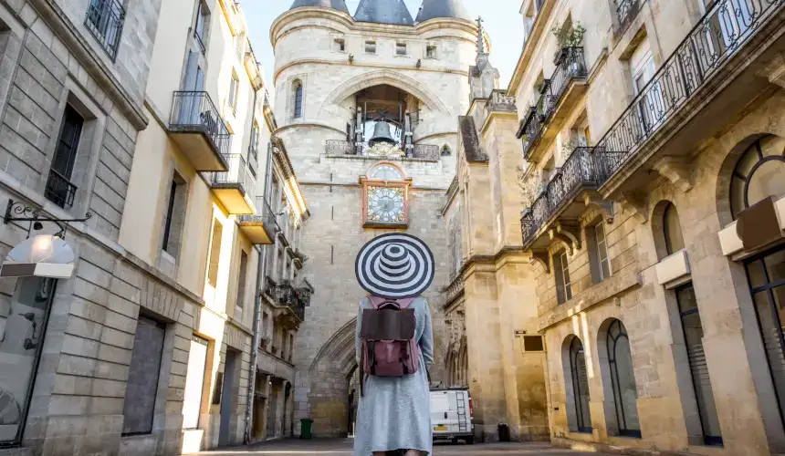 A woman with her back to the camera, wearing a black and white sun hat looks up at Grosse Cloche Tower for its medieval architecture