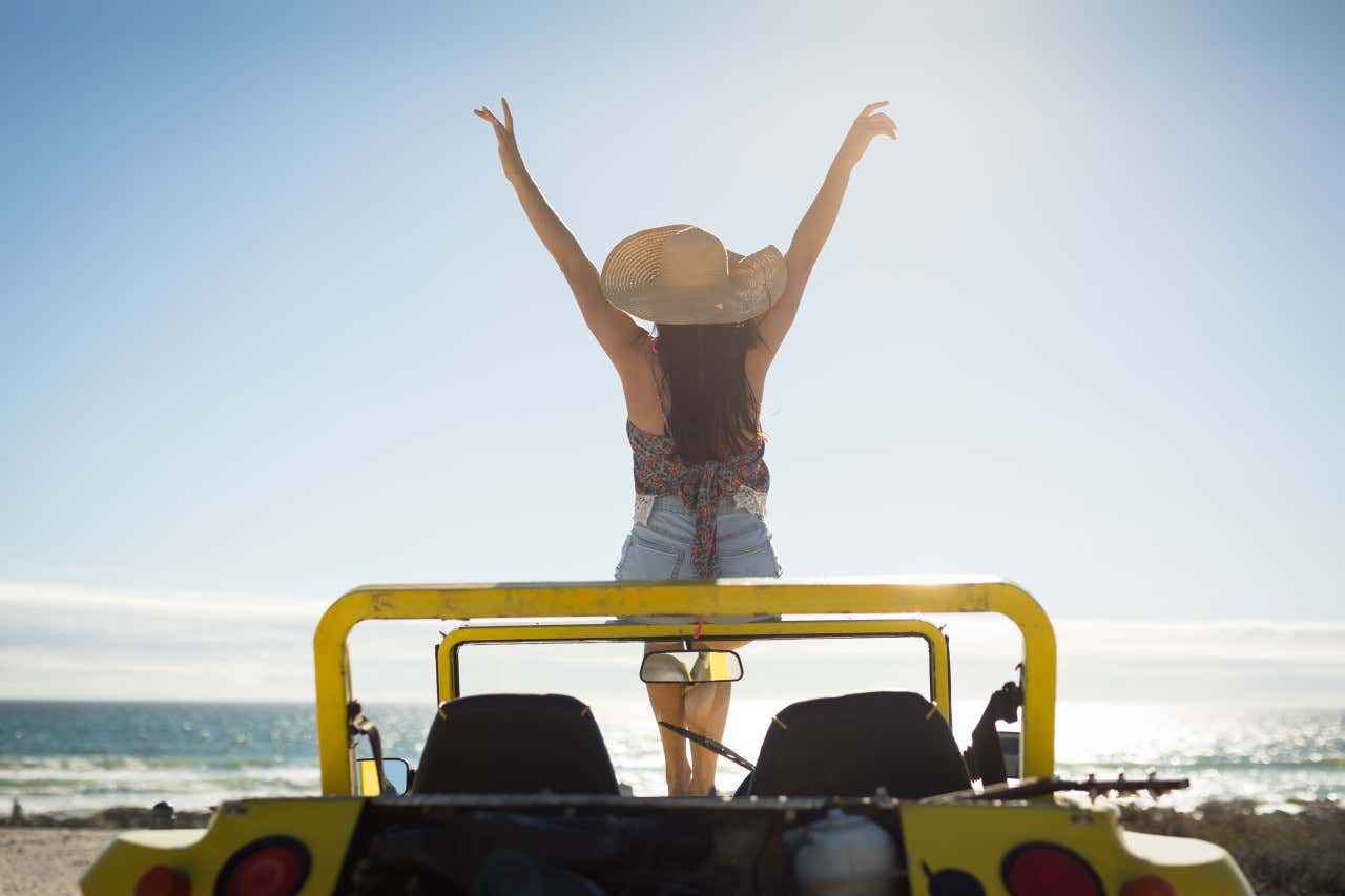 Una mujer de espaldas sentada en la parte delantera de un coche amarillo con los brazos al aire, frente al mar