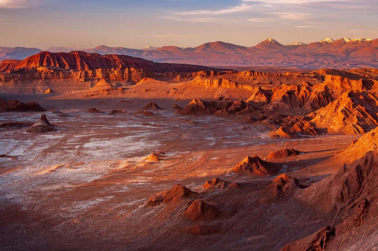 Uma enorme cratera lunar com depósito de sal em meio a um terreno sinistro no Valle de la Luna. A luz do pôr do sol projeta sombras nas montanhas e muda suas cores.