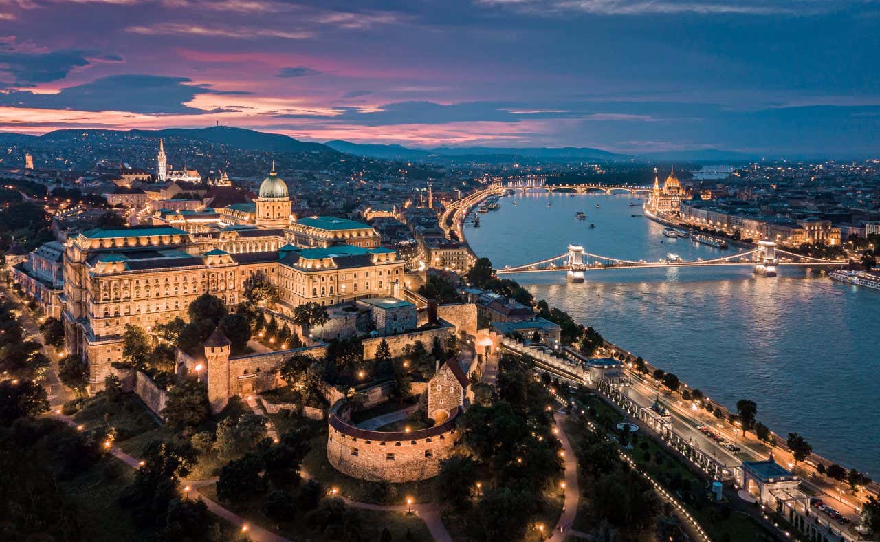 An aerial shot of Buda Castle with the river running along the right at sunset, with the city lit up in lights.