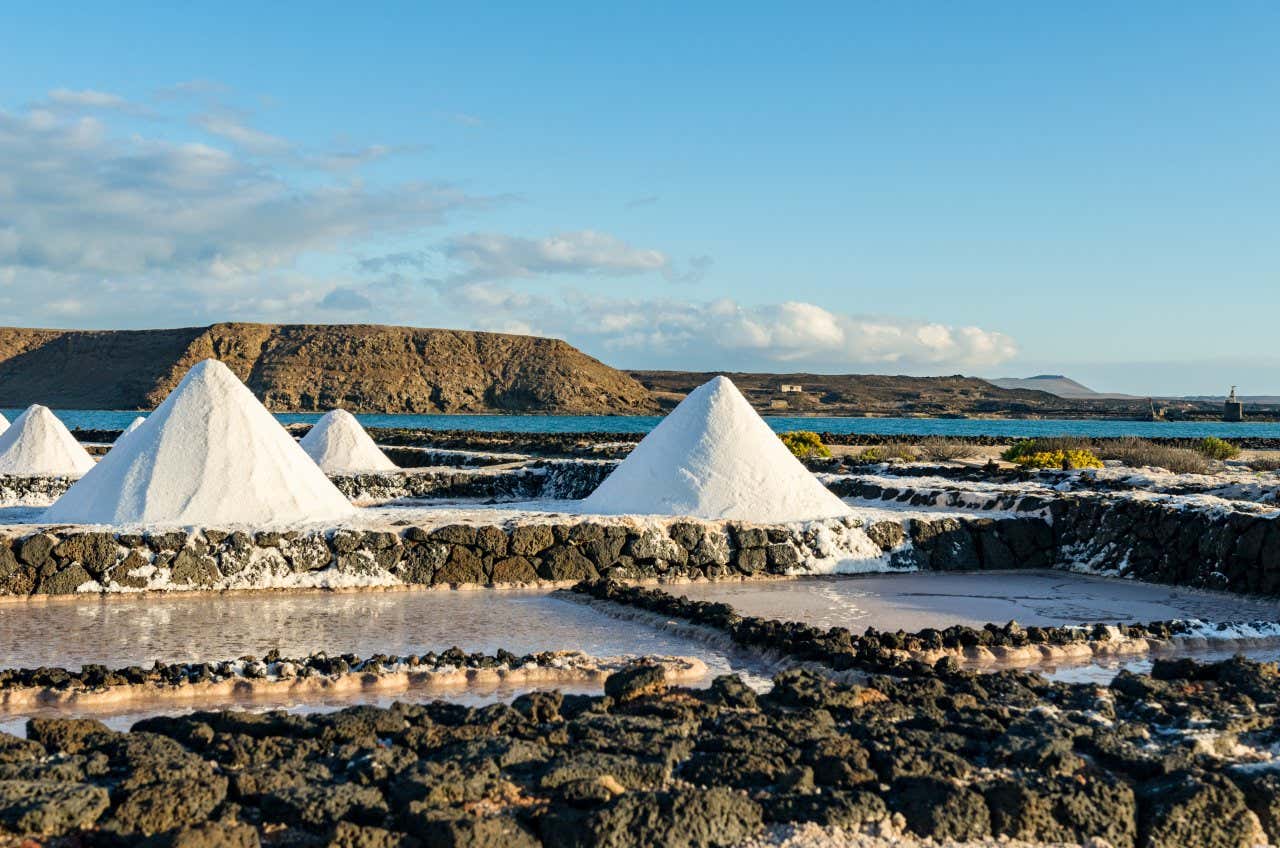 Various heaping mounds of salt at the Janubio Salt Flats in Lanzarote, with the ocean and hills in the background.