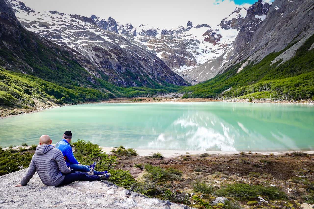 Two tourists contemplating a green lagoon with snow-capped mountains in the background