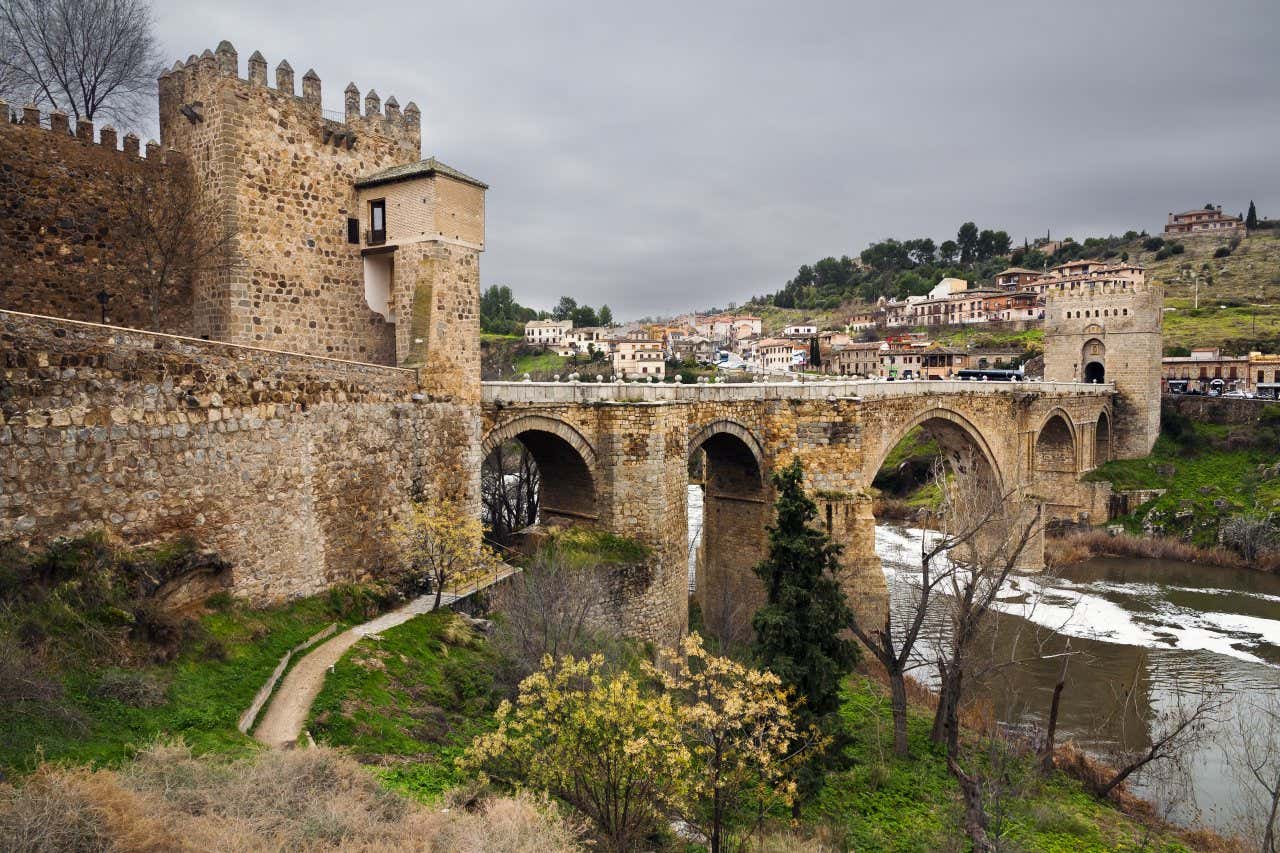 View of the medieval bridge of San Martín and the Tajo river, in Toledo, with an overcast sky in the background.