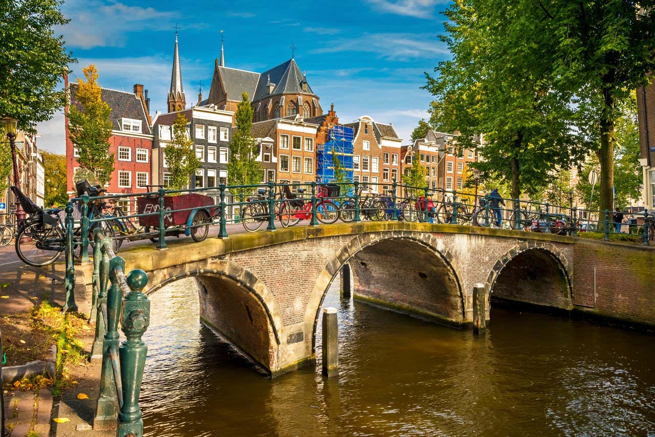 A bridge across the canal in Amsterdam, with bicycles locked to the fence going along the waterside, and buildigns and a cloudy sky visible in the background.