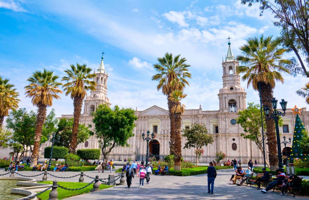 People standing in front of and walking around the Basilica Cathedral of Arequipa, with palm trees in front of it.