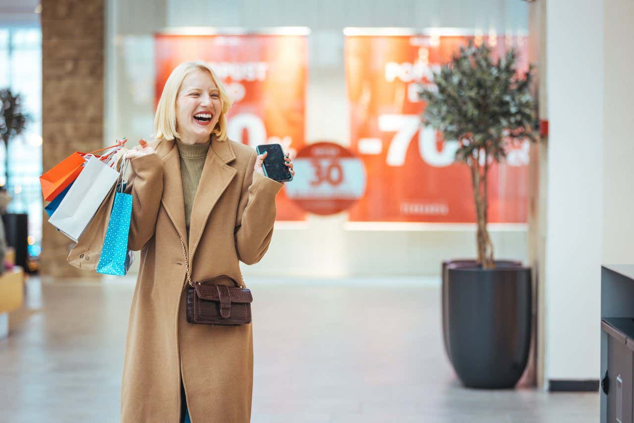 A woman in a long brown coat holding shopping bags in one hand and her phone in another.