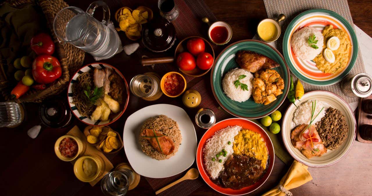 Several plates of Peruvian food on a table with a jug of water, seasoning and other ingredients on a wooden table.