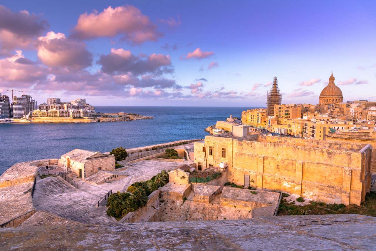 The coast of Valletta, with brick buildings in the foreground, and modern buildings seen across the water, and a cloudy sky above.