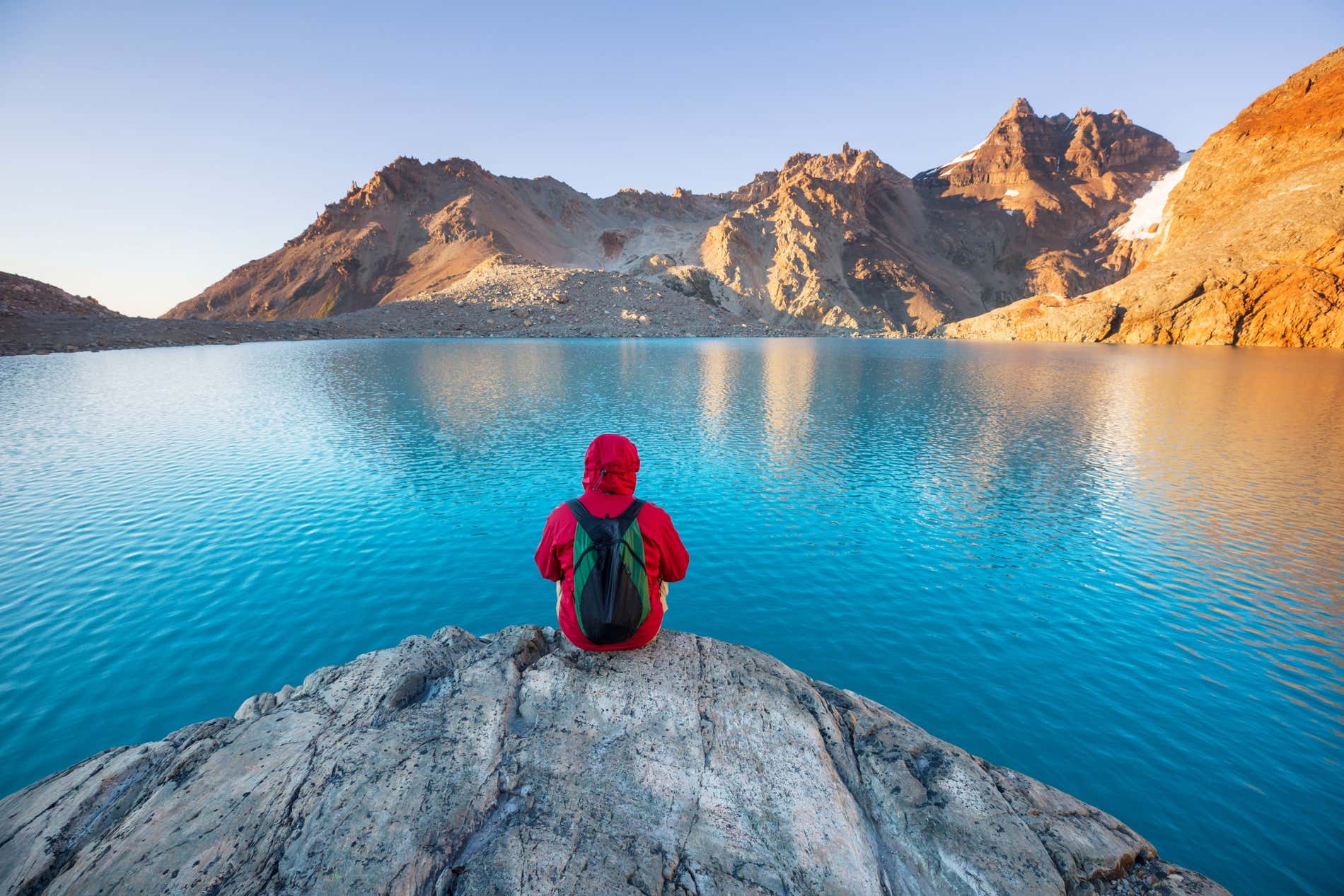 Homem sentado de costas em uma pedra admirando um lago azul com montanhas ao fundo