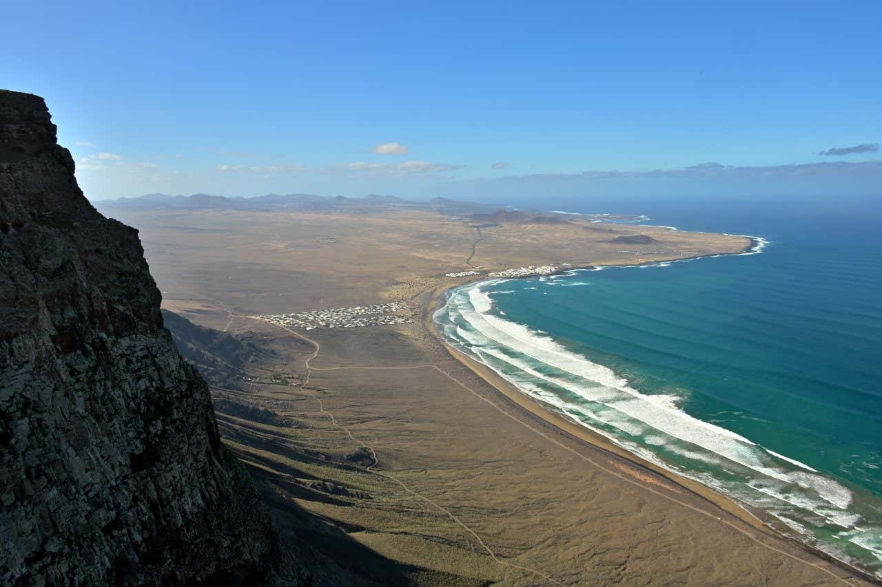 Aerial view of Famara Beach, with waves coming in, and a small town in the distance.