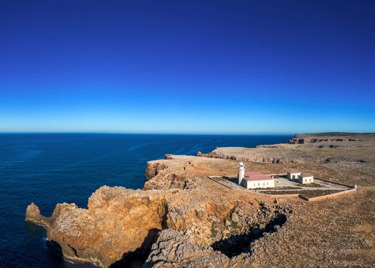 An aerial view Punta Nati Lighthouse as seen from afar, with the sea visible below and the clear sky above.