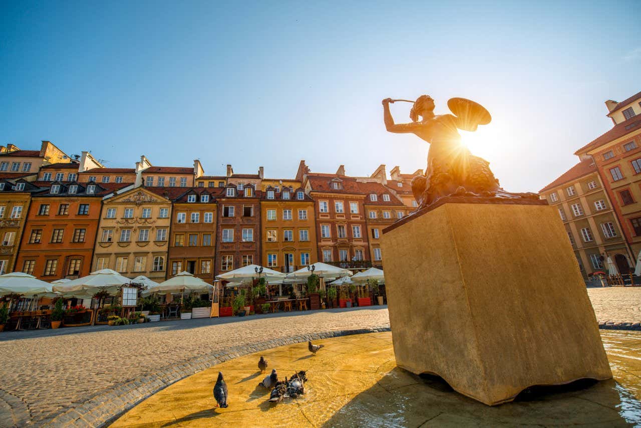 Market Square in Warsaw, with the armed mermaid statue in the middle of the photo, with the sun shining behind it.