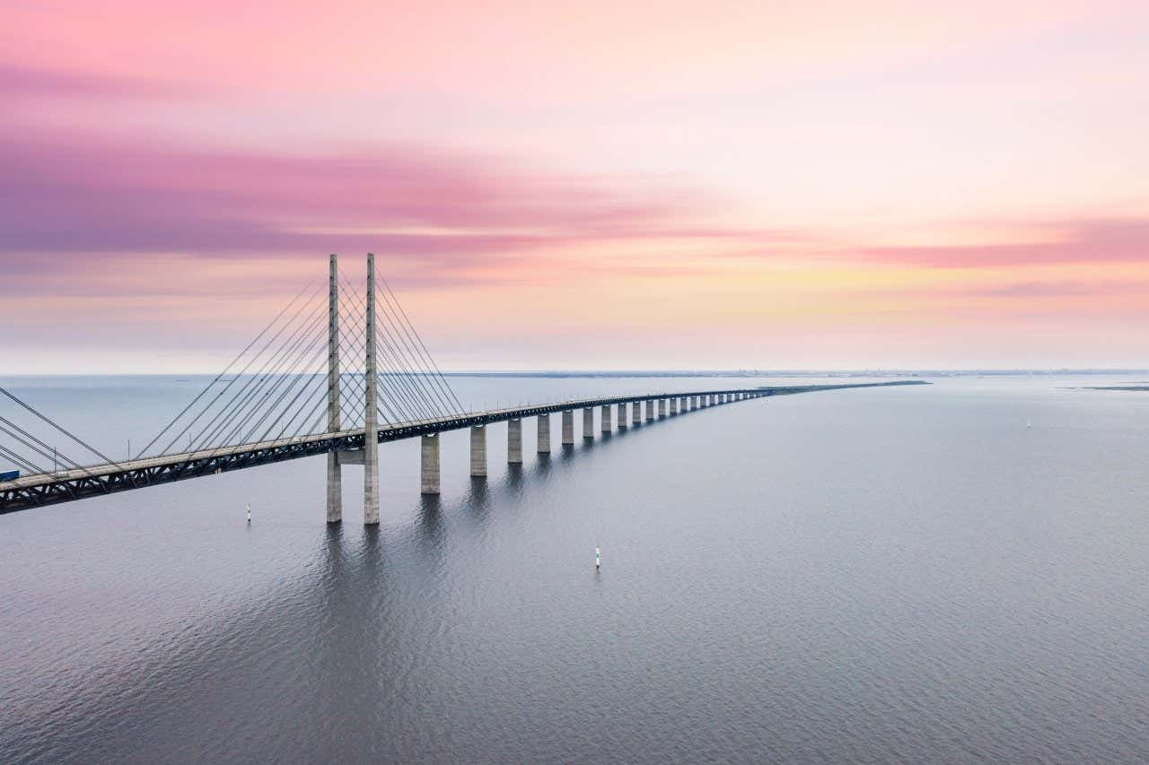 A long grey bridge over the sea leading into an underwater tunnel, under a pink and yellow sky.