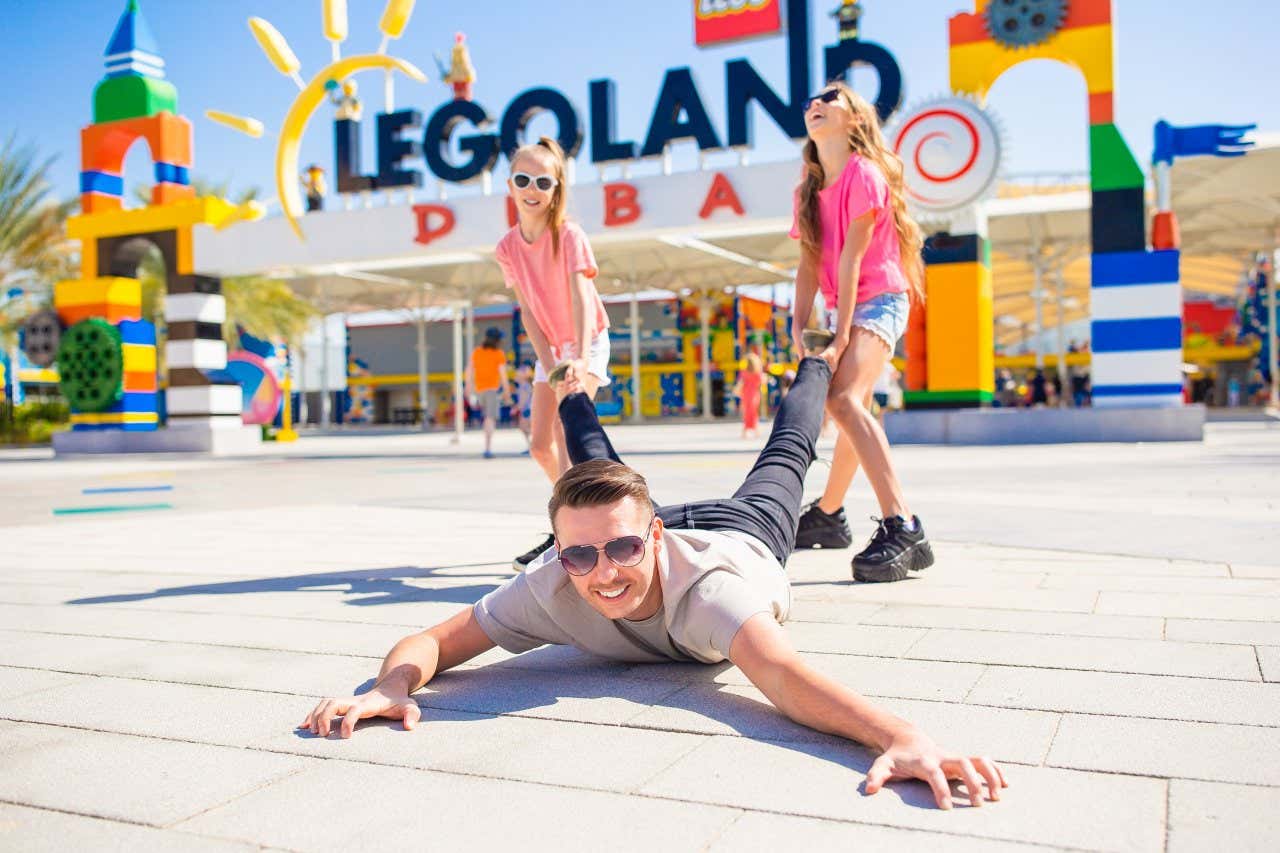 Deux filles vêtues d'un t-shirt rose et d'un short en jean tirant les jambes d'un homme allongé sur le ventre à l'extérieur de LegoLand Dubaï