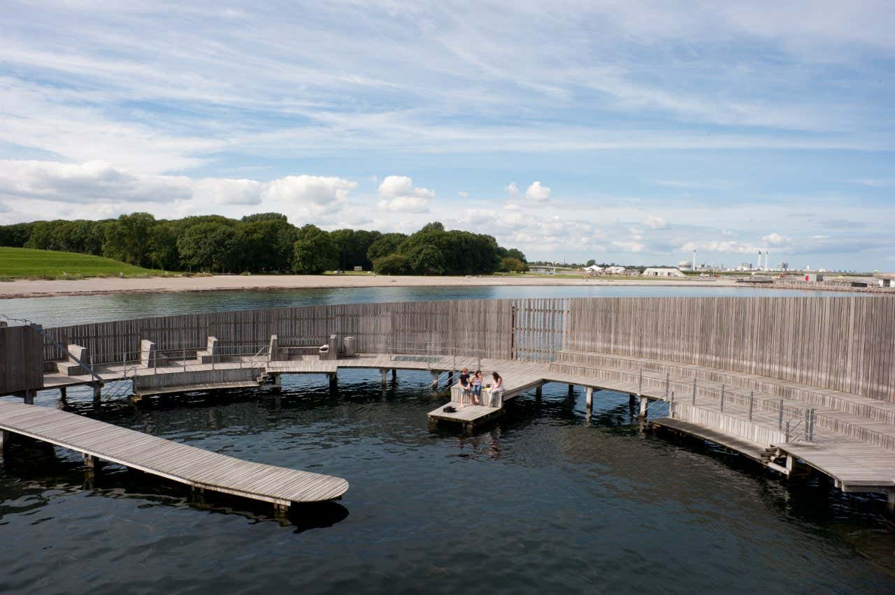 Three people sat on the edge of a wooden jetty around a natural pool in Copenhagen.