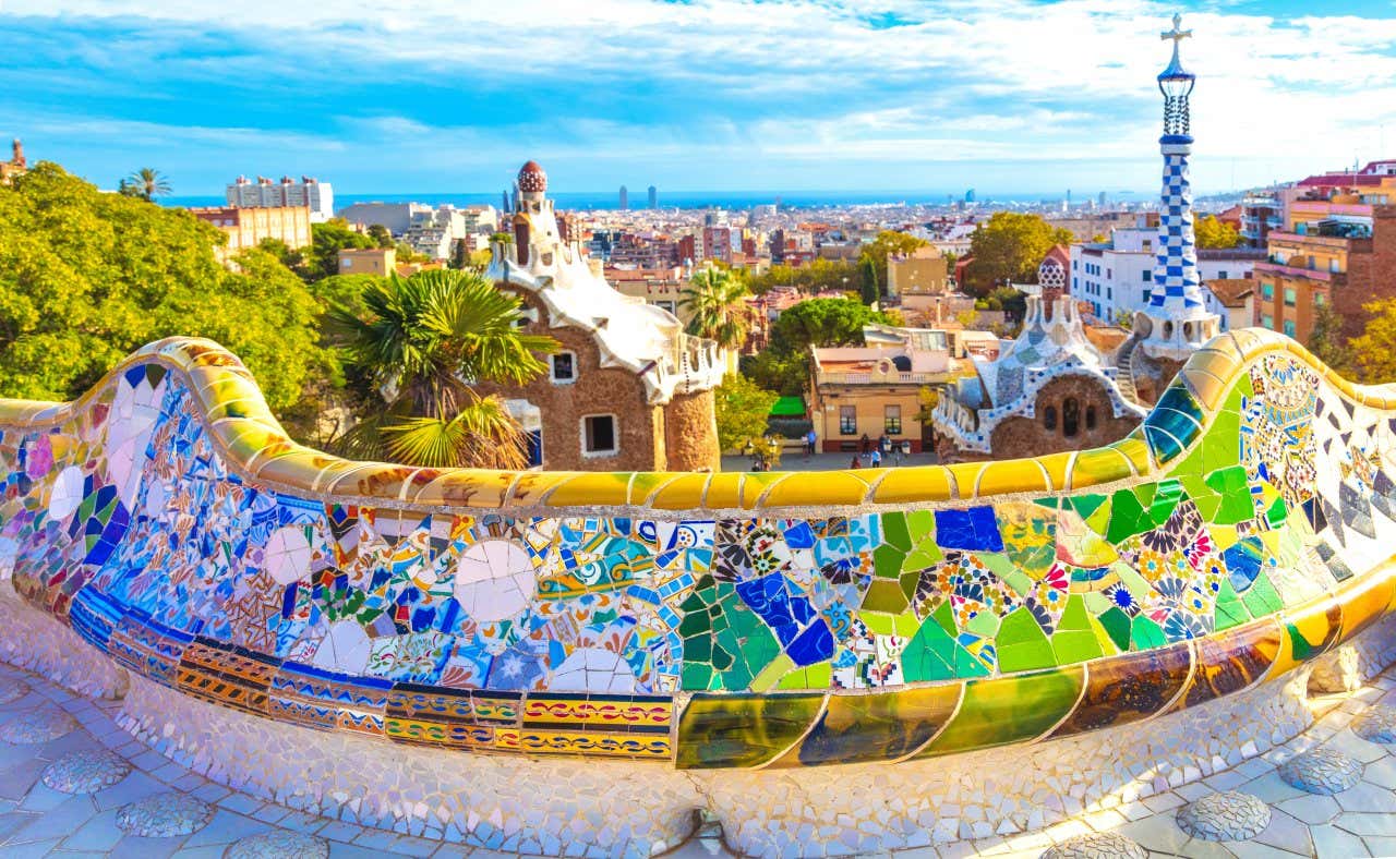 Un muret décoré de mosaïques colorées surplombant des bâtiments en pierre aux toits couverts de mosaïques et la ville de Barcelone