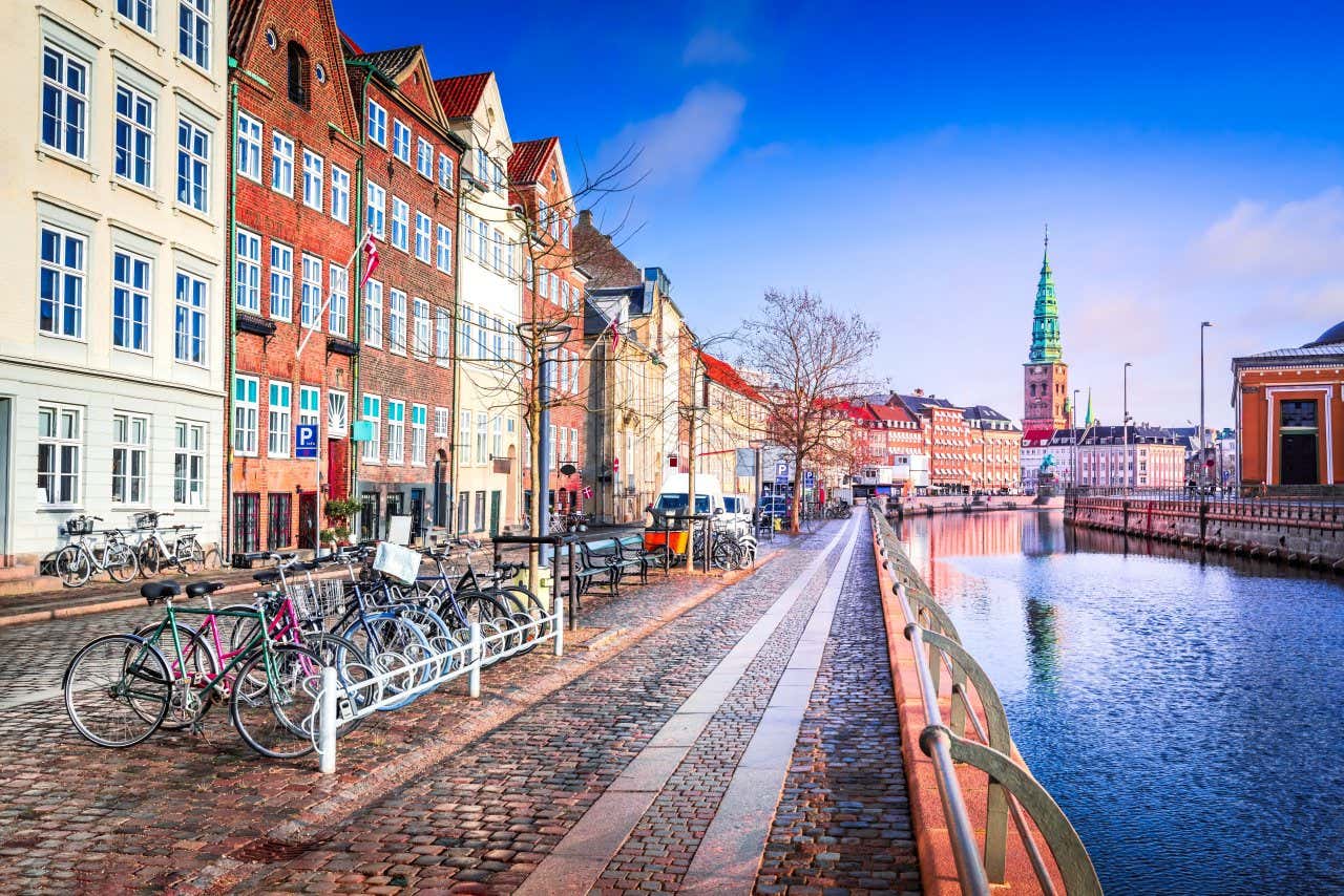 Tall terraced buildings along a cobbled road with parked bikes by a canal on a bright day.