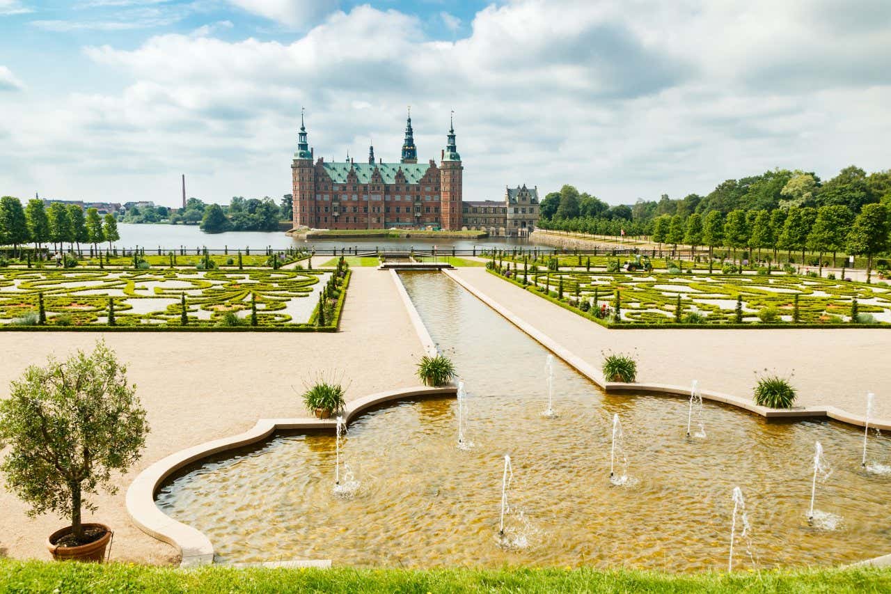 A red brick castle with a copper roof behind a lake and neat gardens and water features.