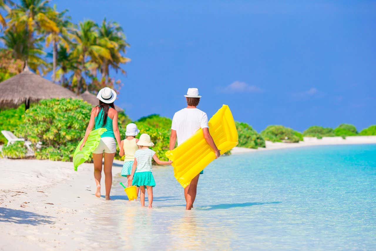 Deux adultes et deux enfants se tenant par la main alors qu'ils marchent sur une plage de sable blanc au bord d'une mer en tenant des flotteurs