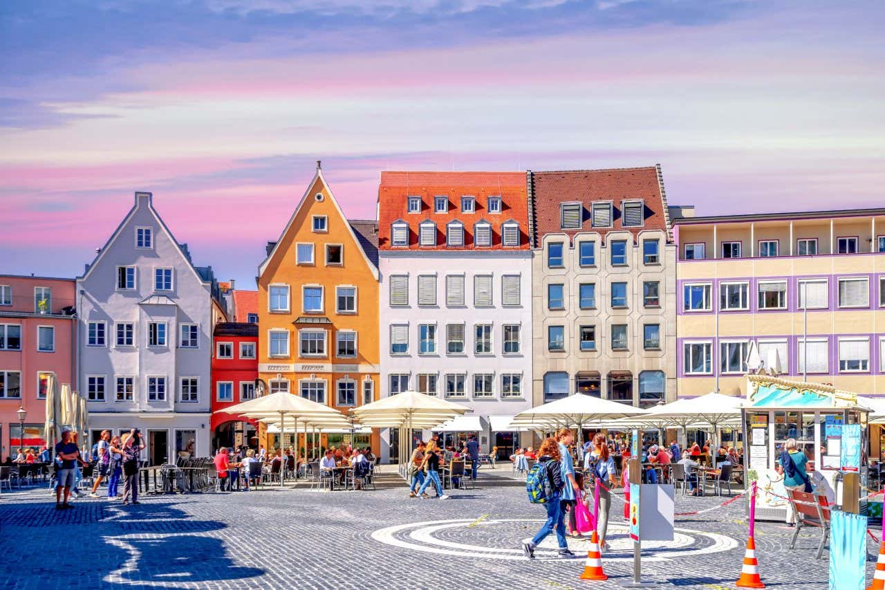 Praça no centro histórico de Augsburgo com chão de pedras e edifícios históricos e coloridos ao fundo com muitas pessoas peasseando 