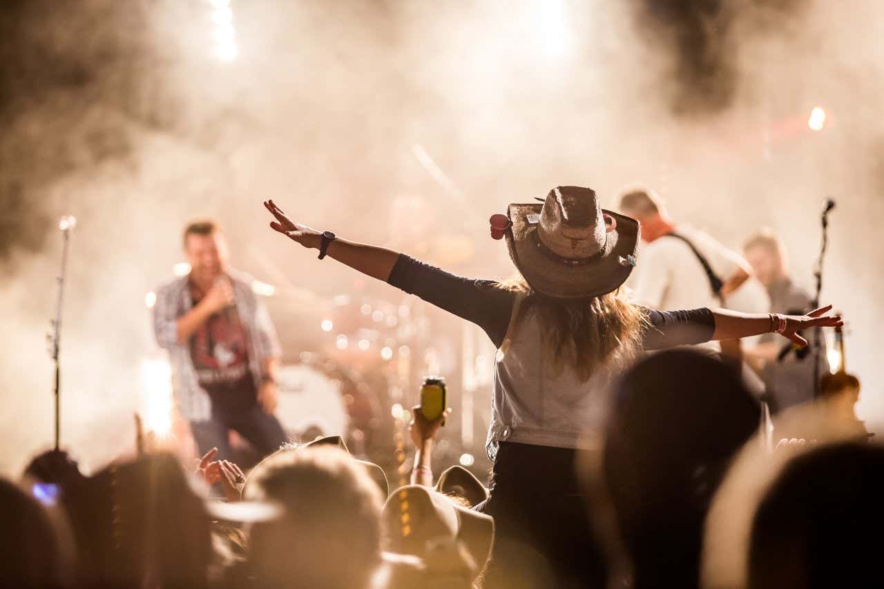 A woman wearing a cowboy hat holding out her arms and standing in a corwd enjoying a live music show.