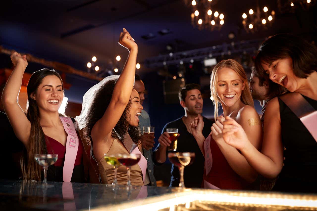 A group of girls wearing pink sashes dancing in a night club near the bar.