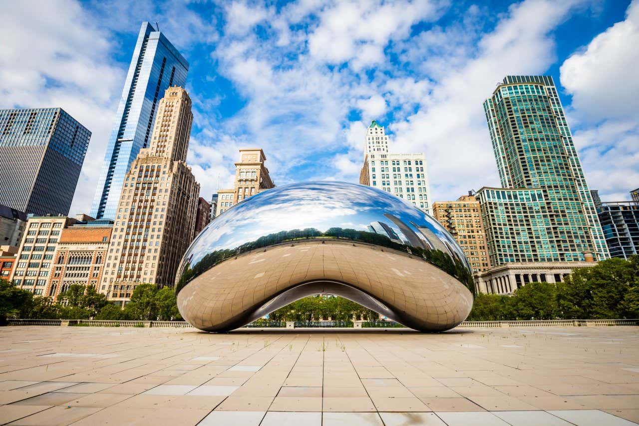 A escultura The Bean no Millennium Park em Chicago, em um dia ensolarado, com arranha-céus e outros edifícios atrás dela, a superfície metálica refletindo o parque em frente