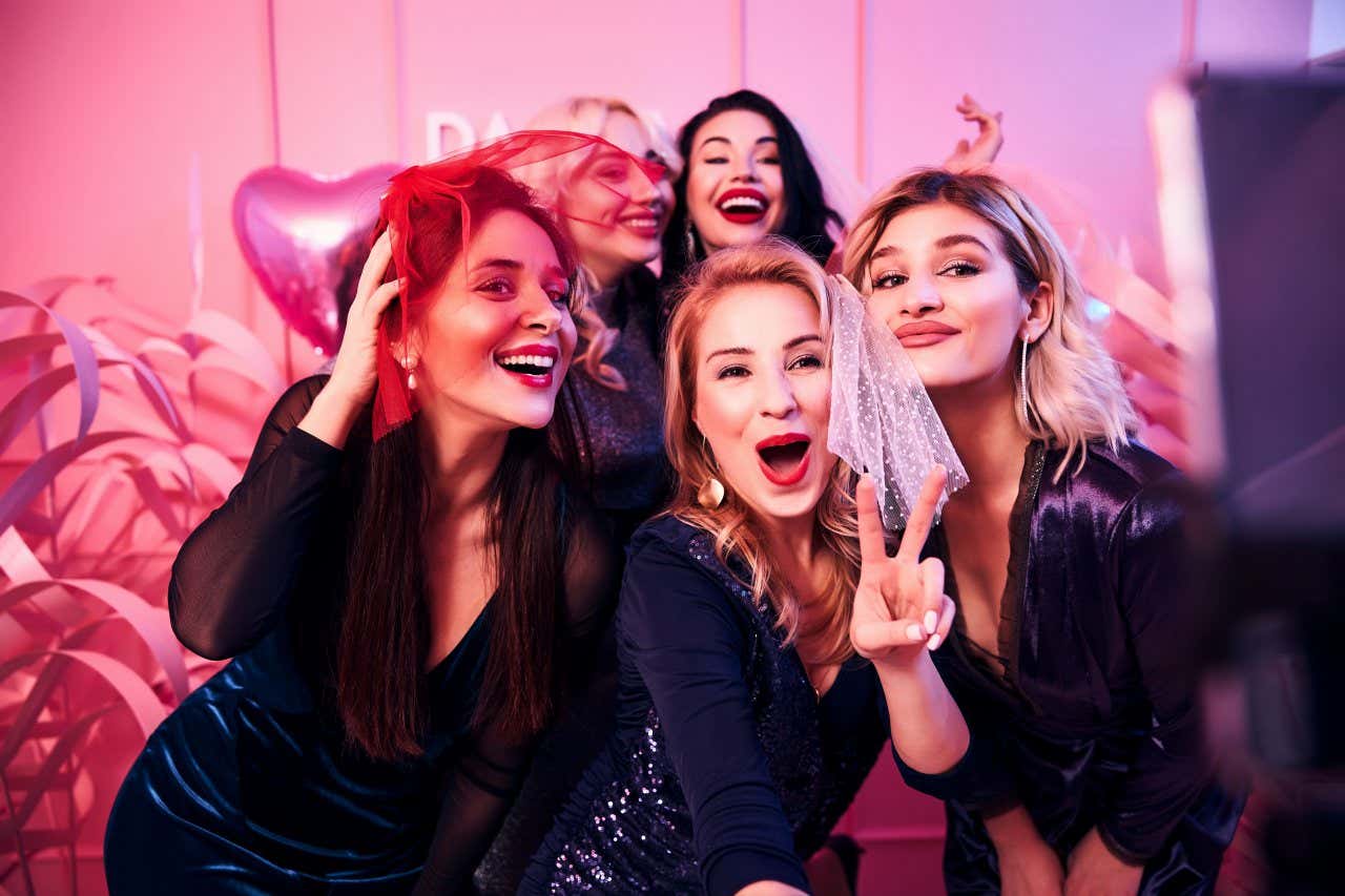 5 women wearing black dresses and red lipstick, with the girl in the middle wearing a white veil and another wearing a red one, posing for a selfie.