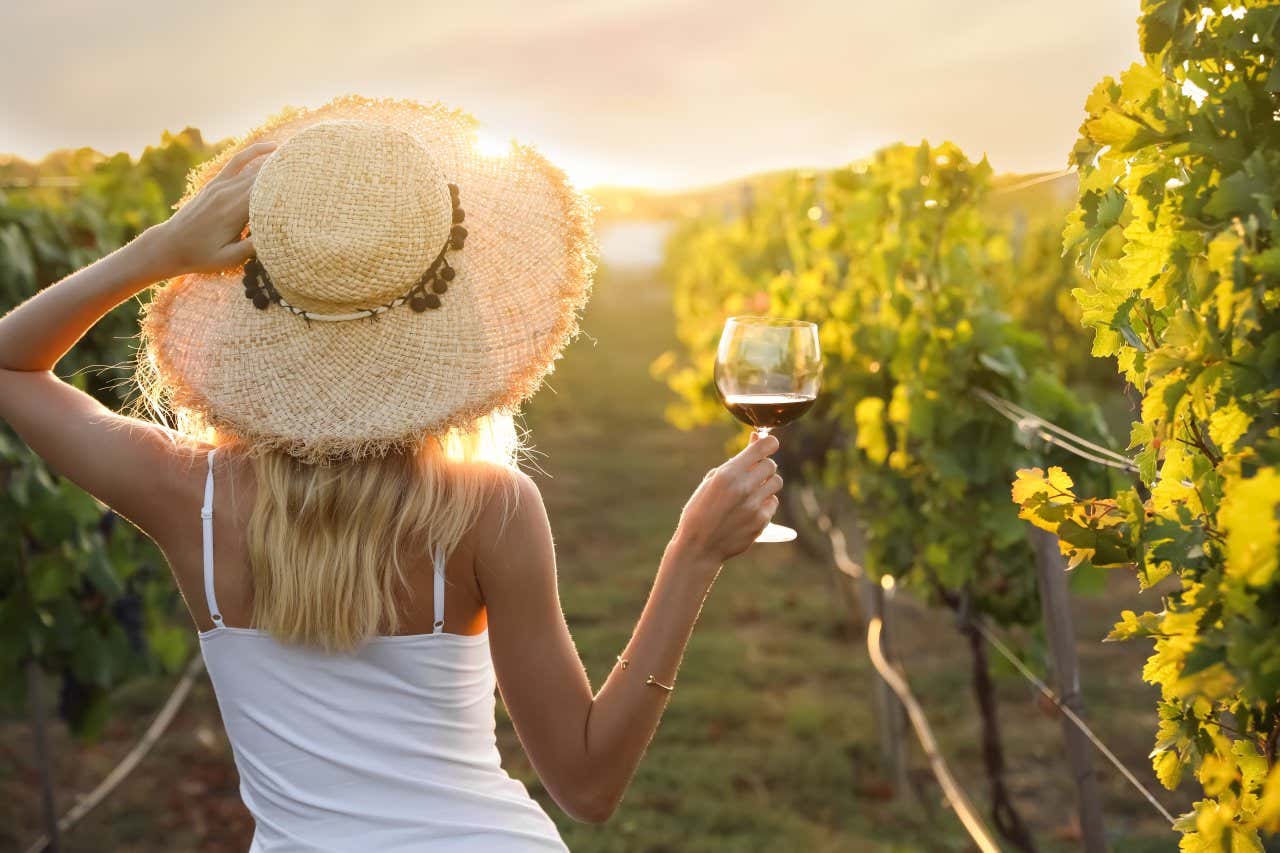 A woman in a white top and a straw hat holding a glass of wine in a vineyard.