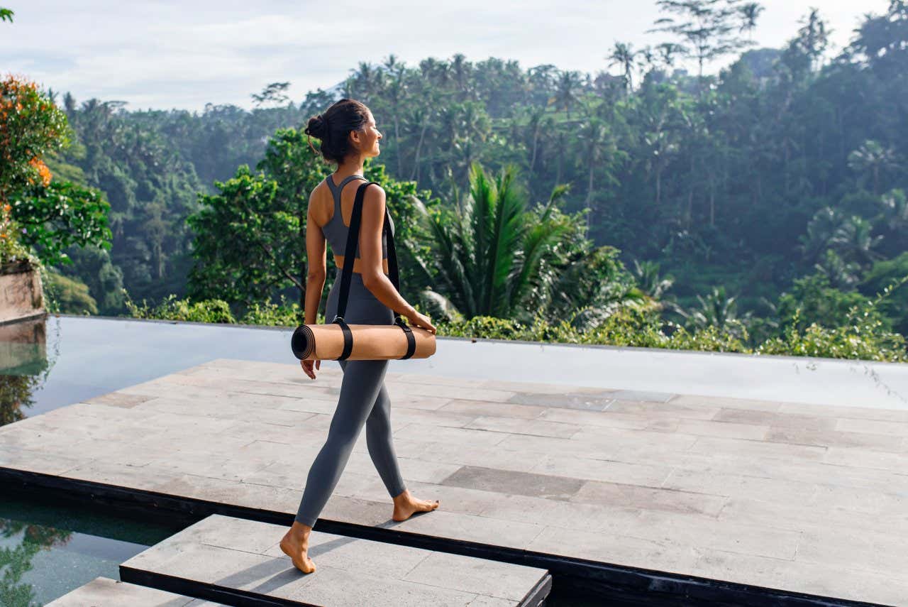 A woman walking with a yoga mat in Ubud, Bali, with a forest visible in the background.