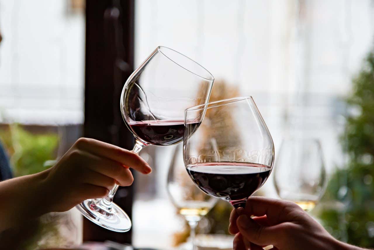 Primo piano di 2 mani con due bicchieri di vino rosso che brindano, con uno sfondo sfocato.