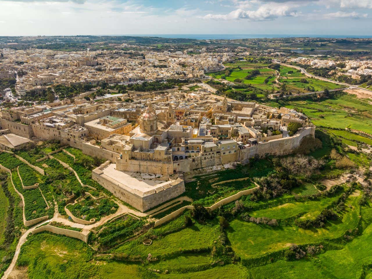 Aerial view of the small fortified medieval town of Mdina with domes and towers surrounded by green countryside.