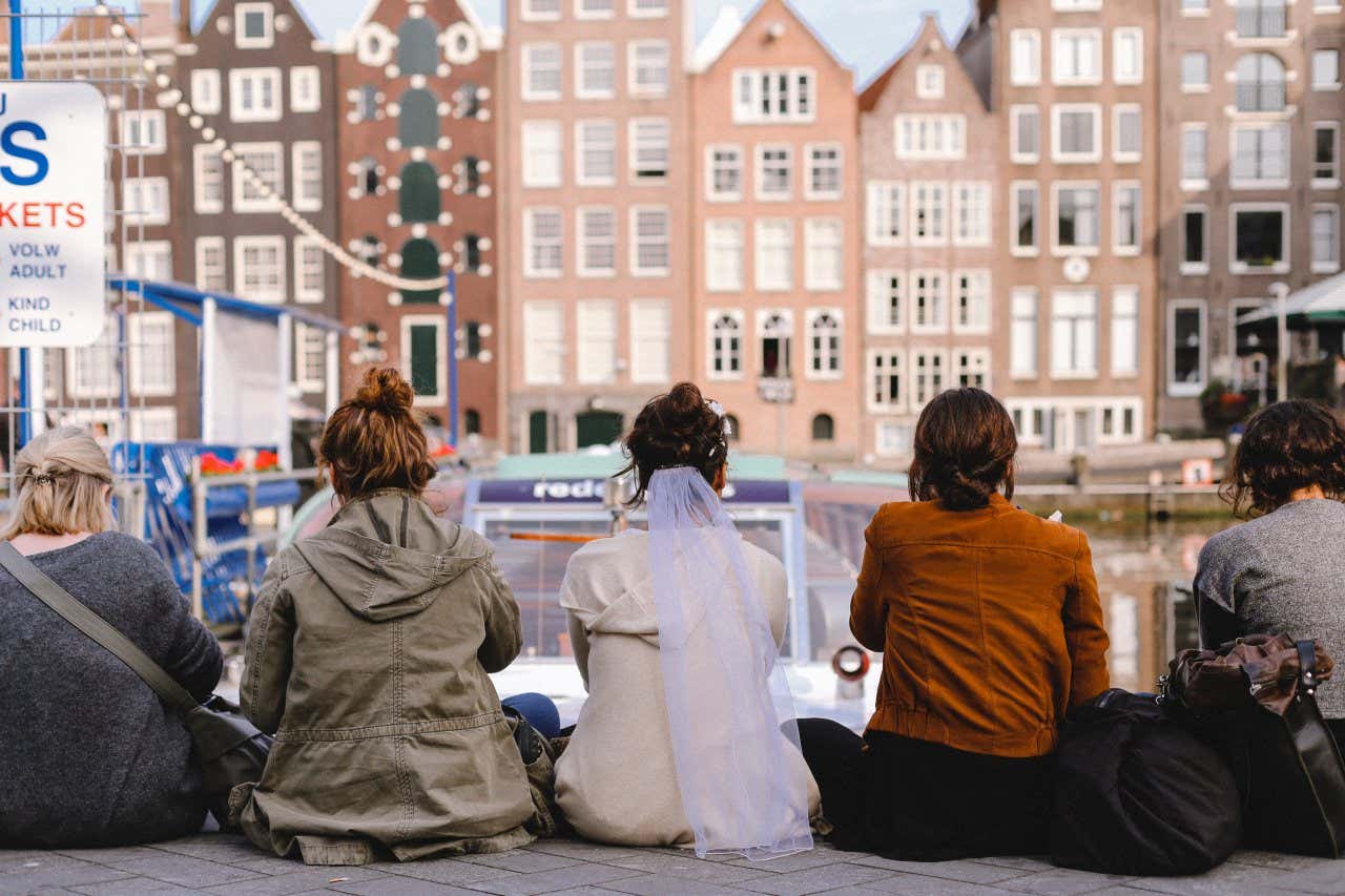 5 women sitting at the edge of a canal, with the woman in the middle wearing a white veil.