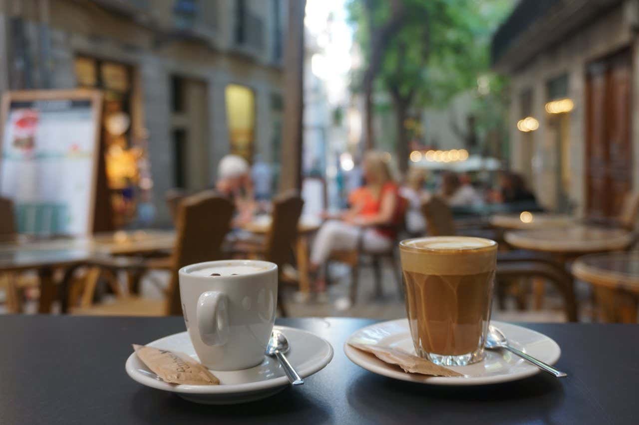 Una tazzina e un bicchiere di caffè affiancati su un tavolo, con uno sfondo sfocato dei tavolini di un bar all'esterno.