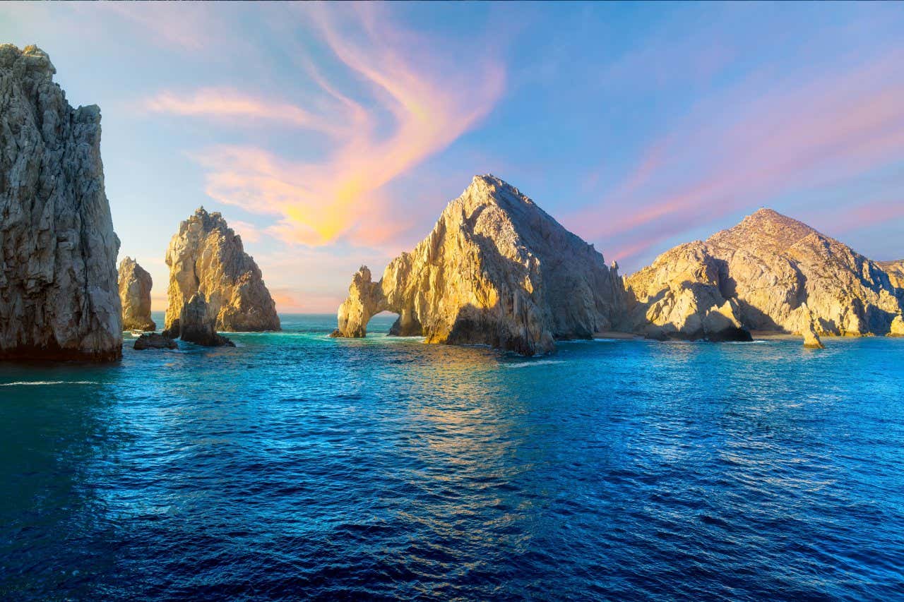 A view of rock formations, and arches in the sea under a blue sky with orange and yellow hues in it.