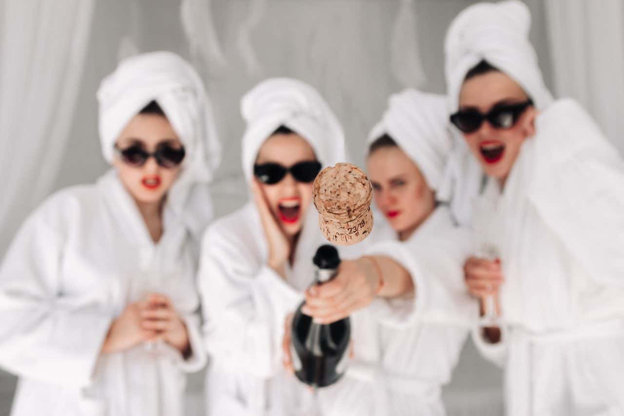 4 women wearing white bathrobes and towels on their heads with black sunglasses and red lipstick popping a bottle of bubbly towards the camera!