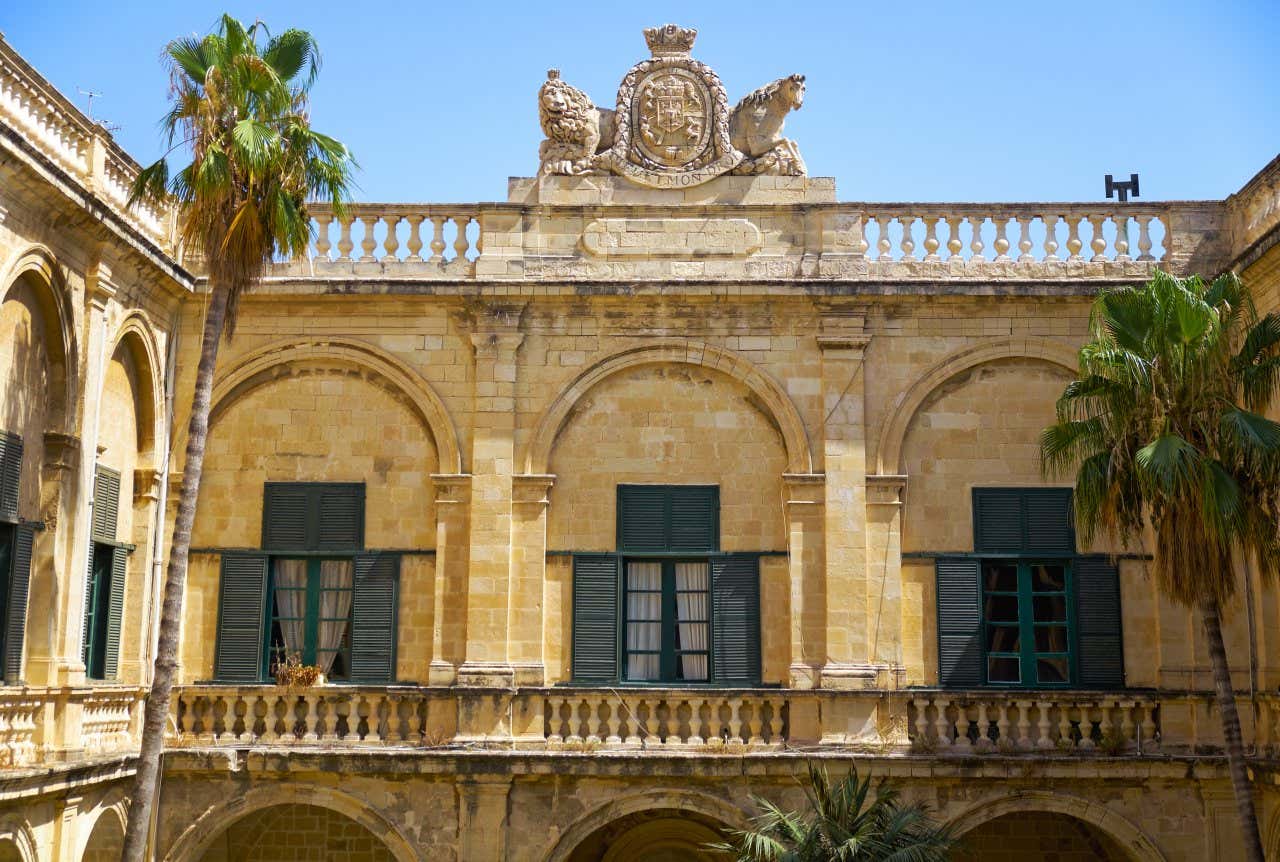 Yellow stone coat of arms with a lion and a horse on either side, surmounting a row of windows inside a patio of an old palace in the centre of Malta.