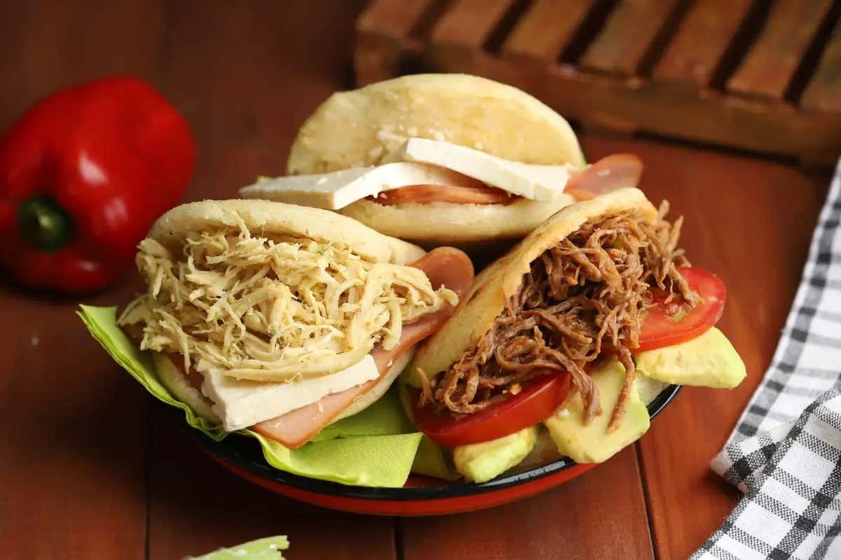 Arepas recheadas com frango, carne e queijo em um prato em cima de uma mesa de madeira com um pimentão vermelho ao lado