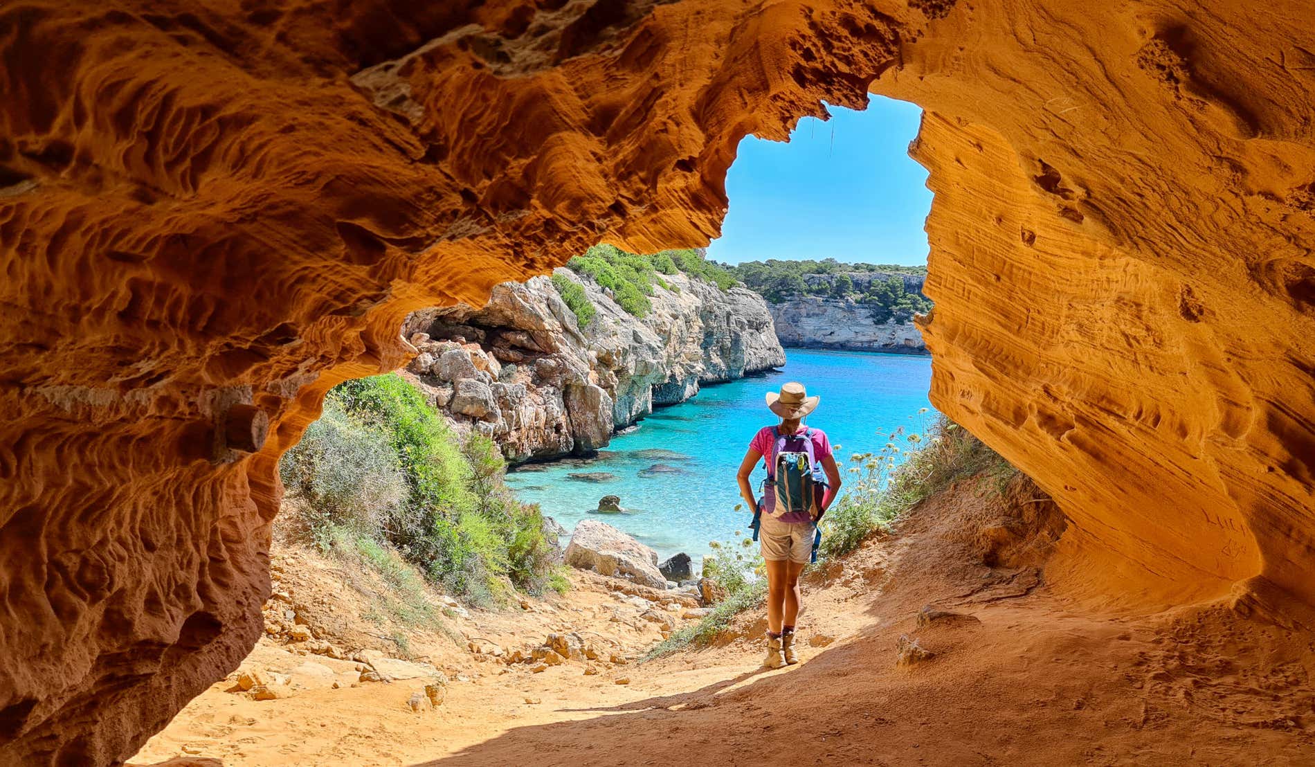 Un turista con sombrero de espaldas a la cámara entre un arco de piedra natural que ofrece vistas a una pequeña cala de aguas cristalinas en Mallorca