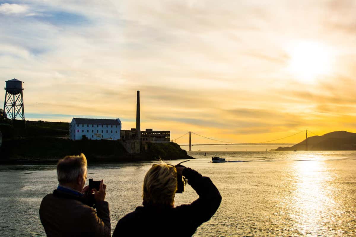 Two tourists on a boat photographing the Golden Gate Bridge at sunset.