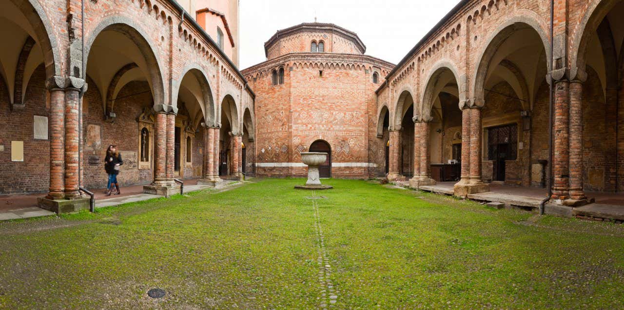 Cortile interno di una chiesa romanica in pietra, sul fondo un edificio in mattoni ottagonale, con una fonte in pietra al centro e due arcate laterali con colonnati