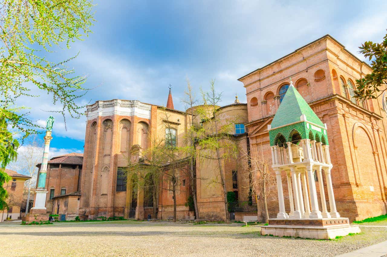 Veduta di una piazza storica a Bologna, con antichi edifici in mattoni e un monumento decorativo in primo piano, il tutto incorniciato da alberi primaverili e un cielo parzialmente nuvoloso