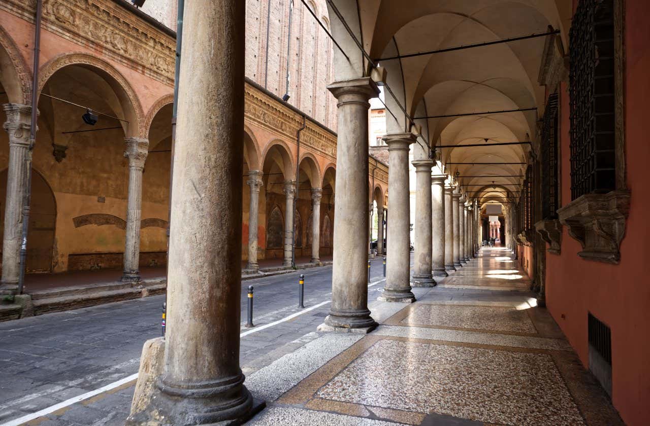 Scorcio di un portico storico a Bologna, con colonne di pietra che si susseguono creando un effetto prospettico, e una pavimentazione in mosaico che riflette la luce del giorno