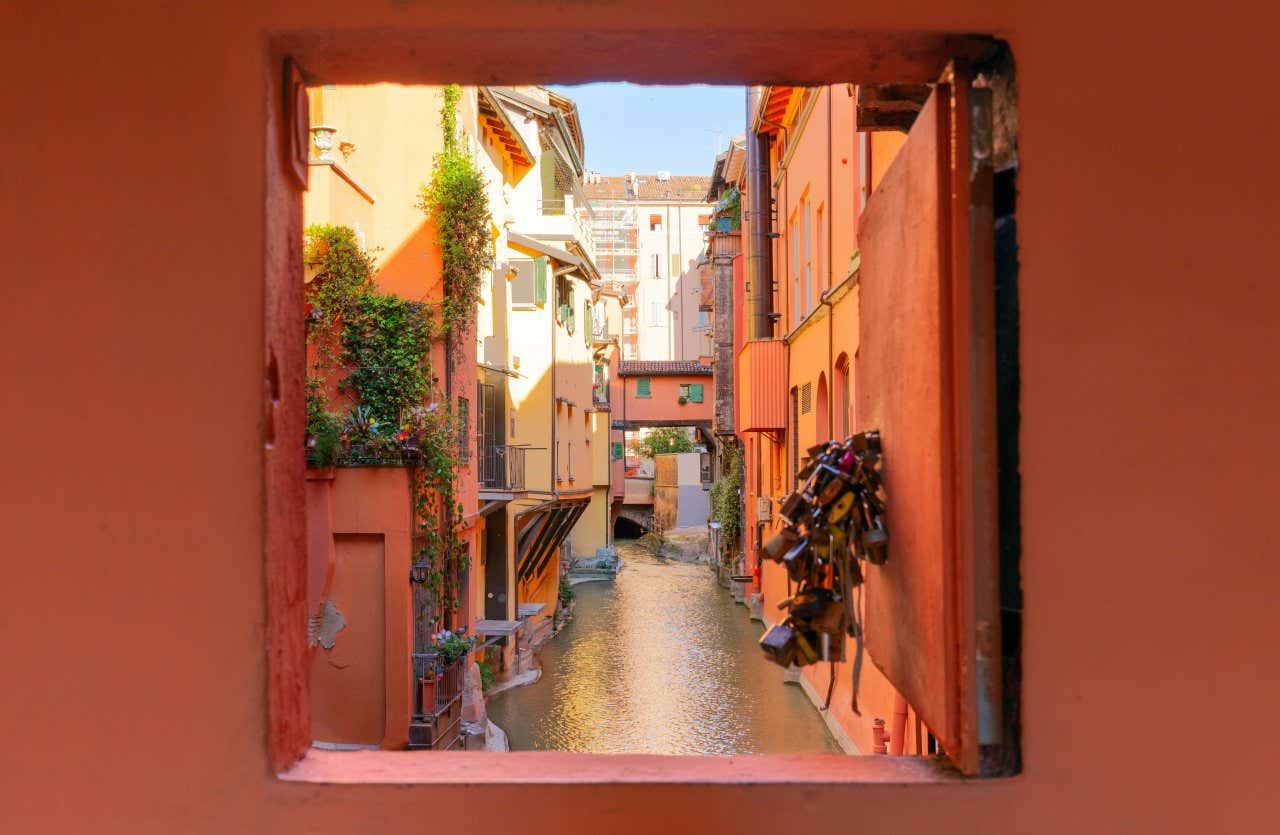 Una piccola finestra rossa aperta con vista su un canale stretto che scorre tra palazzi nel centro di Bologna