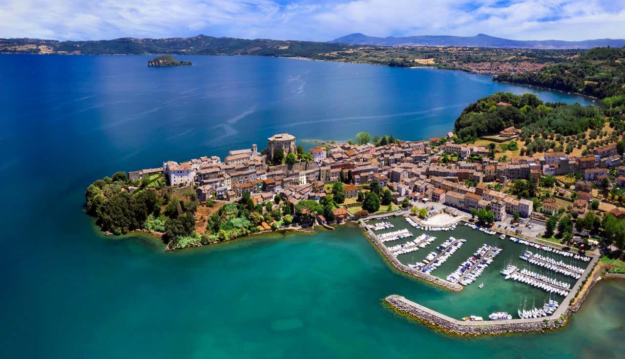 Vista dall'alto del paese medievale Capodimonte affacciato sull'acqua turchese del lago di Bolsena con un piccolo isolotto roccioso in lontananza in una giornata di sole