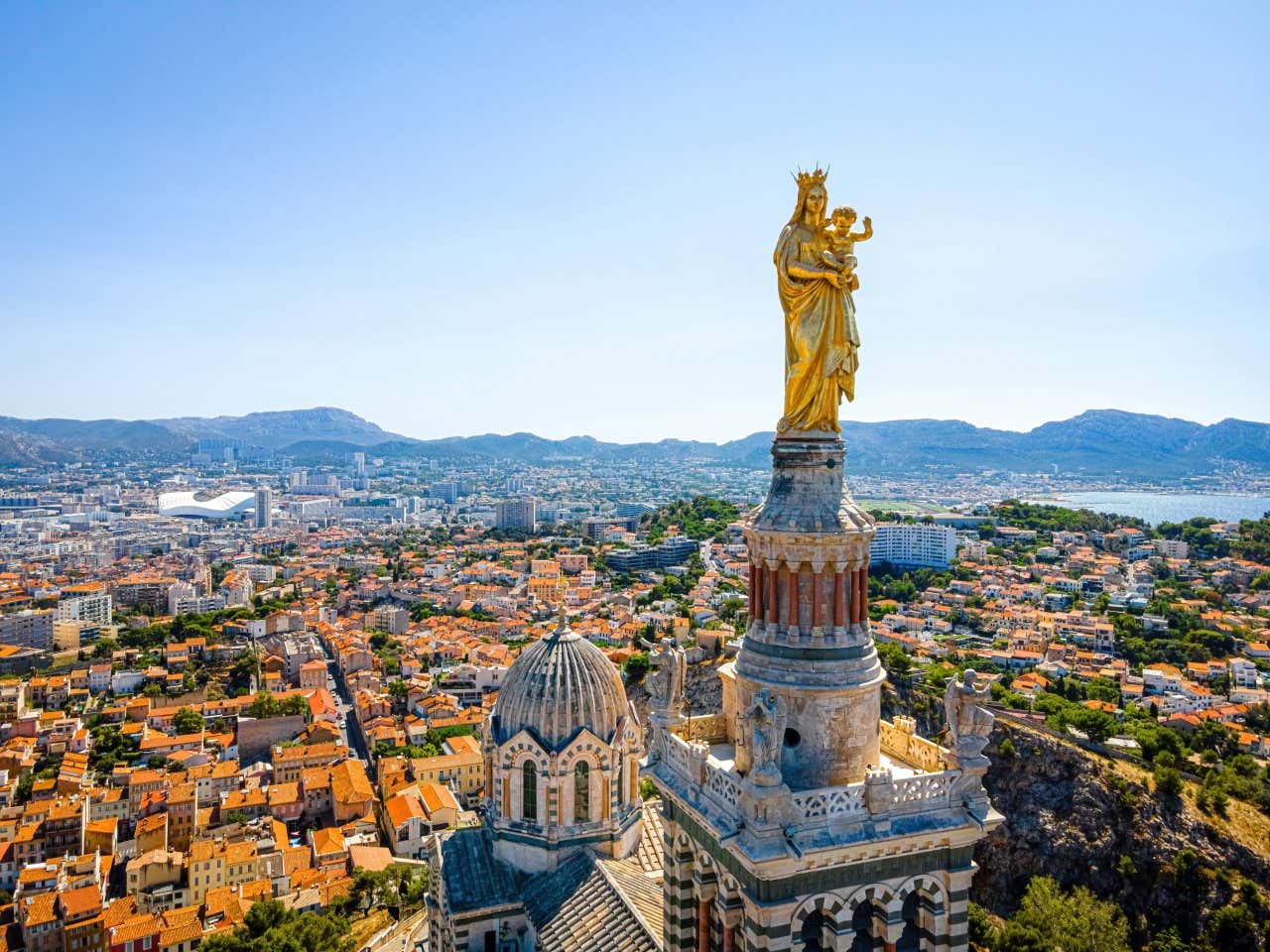 A golden statue of the Virgin Mary and Jesus on top of the basilica with views of Marseille and the coast in the background