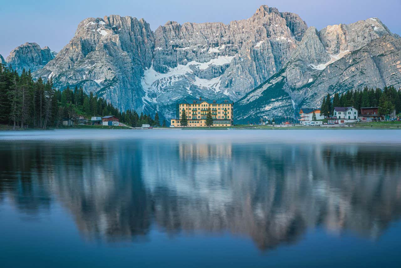 Un grande edificio giallo con tetto spiovente verde alle pendici di grandi montagne innevate e sulle sponde di un grande lago montano