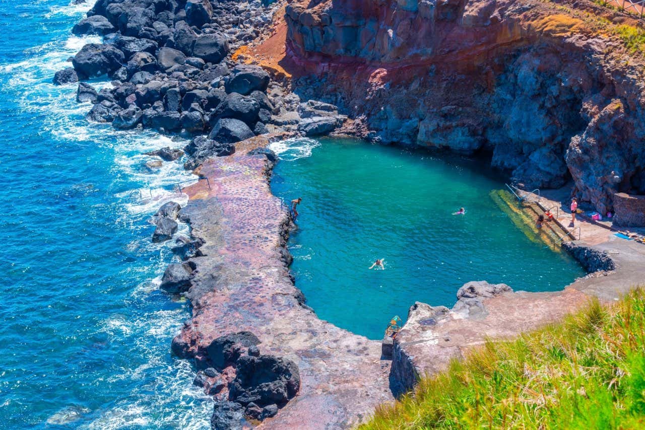 Una piscina naturale di lava sull'isola di São Jorge, con acque di un blu intenso che contrastano con le rocce vulcaniche scure e le pareti rocciose dai toni rossastri. Alcune persone si stanno godendo un bagno rilassante in questo ambiente naturale. La piscina è accessibile attraverso scalette che conducono direttamente nell'acqua.