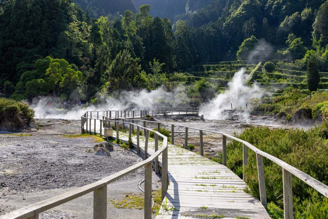 Una passerella in legno attraversa una vallata vulcanica immersa nel verde con colonne di fumo che escono dal sottosuolo